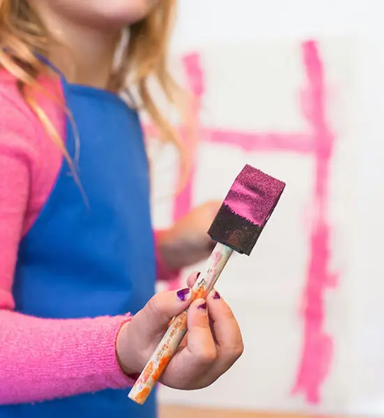 girl-holding-paintbrush.jpg (1) A young girl holding a paintbrush dipped in pink paint.