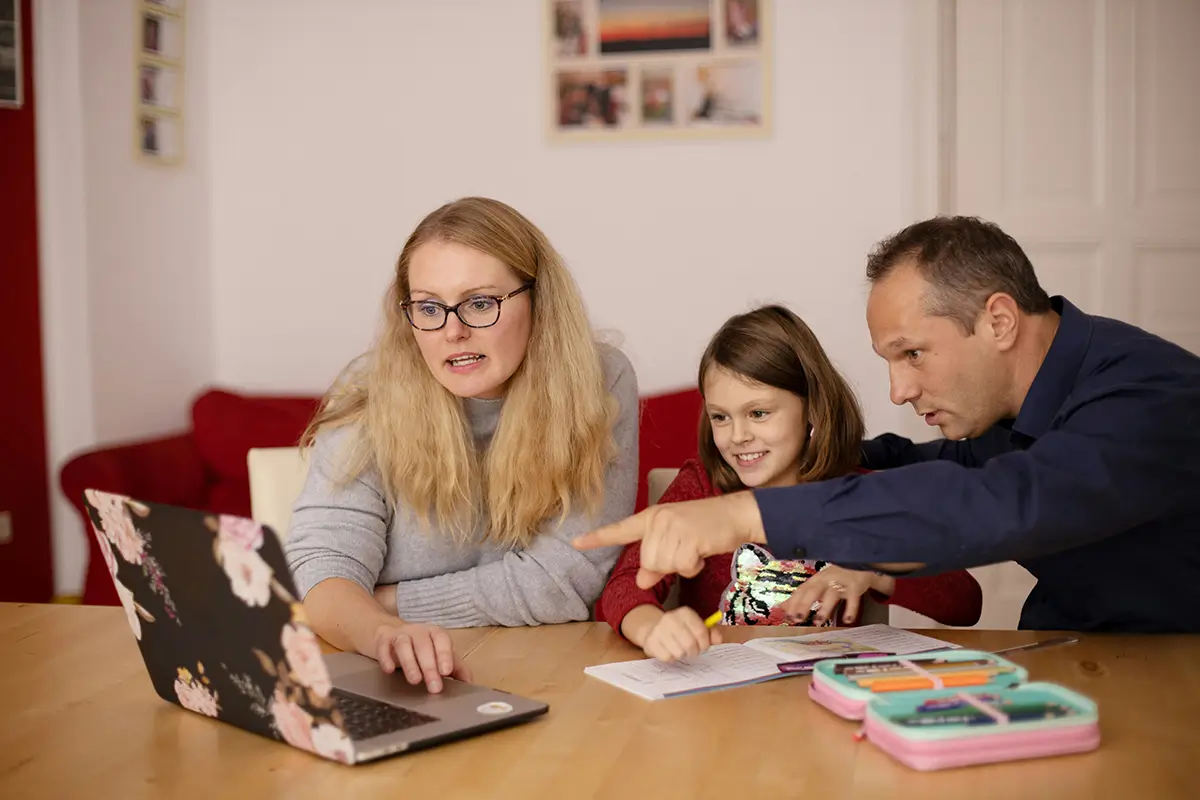 A woman with blonde hair and glasses, a young girl, and a man sit at a wooden table looking at a floral-patterned laptop. The man points toward the screen.