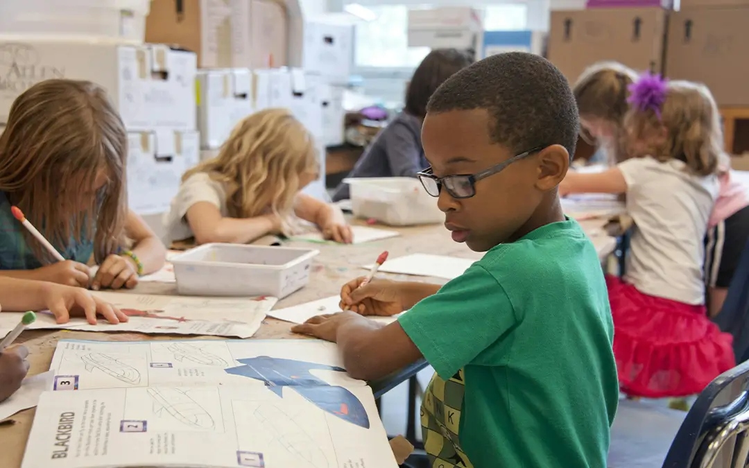 A young boy wearing glasses and a green shirt focuses on drawing in a workbook at a classroom table with other children.